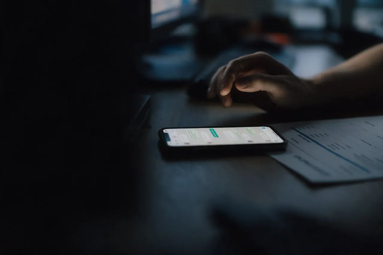 Celular com uma conversa parada sobre a mesa, ao lado de uma proposta de preço impressa, e uma mão que não chega a digitar